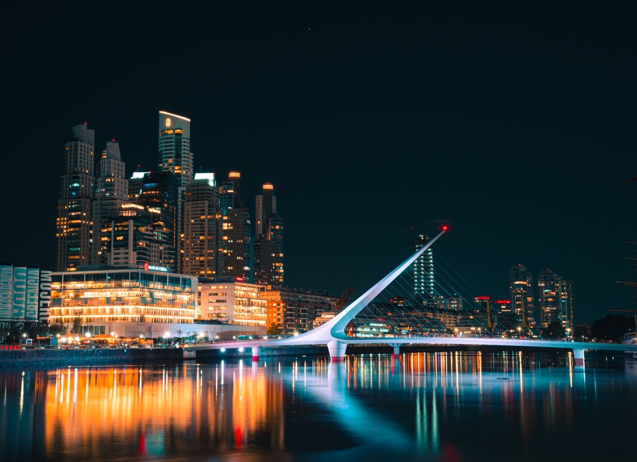 Modern architectural view of Puente de la Mujer and Buenos Aires skyline reflected in water at night.