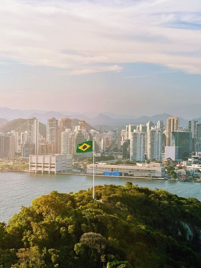 Aerial view of Brazilian flag atop green hill with city skyline in the background.